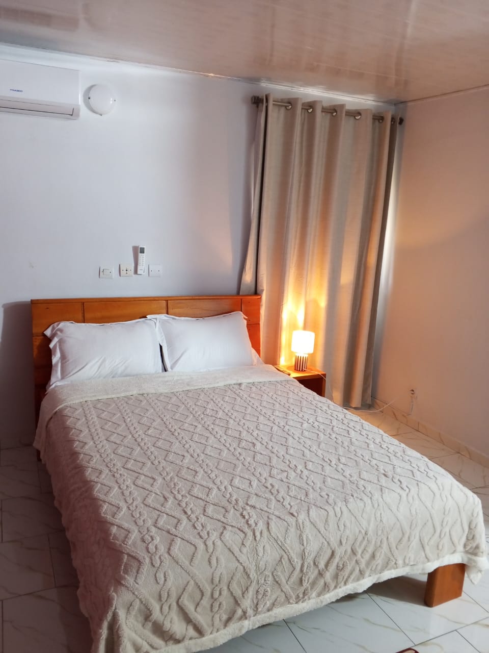Chambre climatisée Békan Tié avec lit double, lampe d'ambiance et rideaux beige